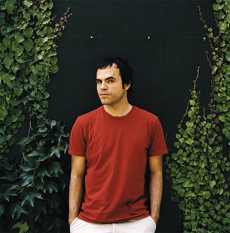 Hossein Derakhshan in a red T-shirt and white pants, standing in front of a black wall with leafy vines running vertically on either side.