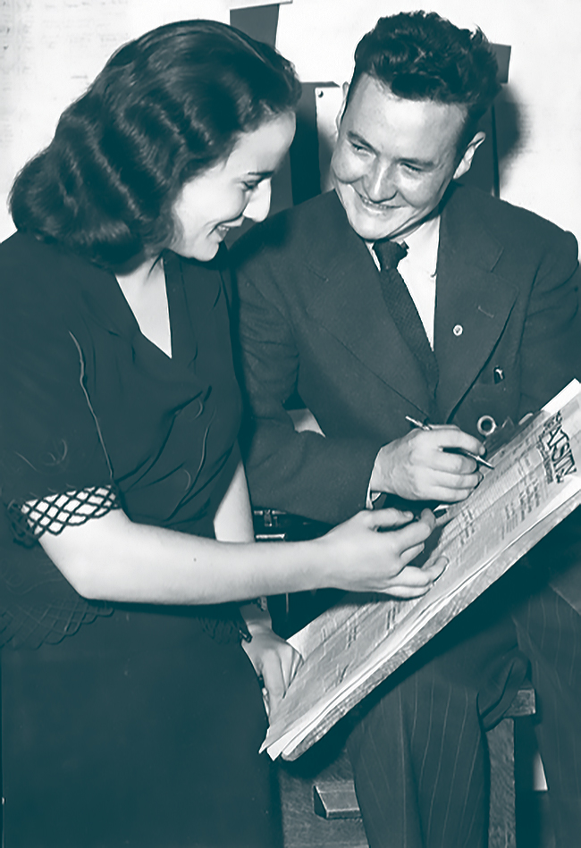 Black and white photo of Betsy Mosbaugh (left) reviewing the pages of a newspaper that Bob Grosskurth is holding out. Grosskurth is looking at Mosbaugh with a pen in his hand.