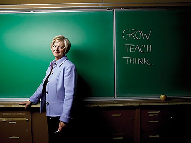 Professor Carol Rolheiser, in a lavender jacket, standing in front of a green chalkboard, holding a piece of chalk in one hand. On the chalkboard are the words "grow," "teach" and "think" in capital letters, each on its own row.