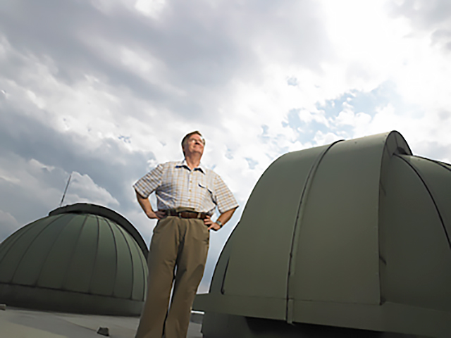 Professor John Percy is standing with his hands on his hips, on a rooftop, next to grey spherical structures.