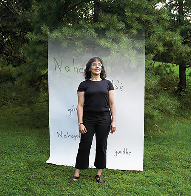 Professor Keren Rice, wearing a black T-shirt and pants, is standing on a grassy field with evergreen trees behind her. Superimposed behind her is a paper with a few words written in Aboriginal language.