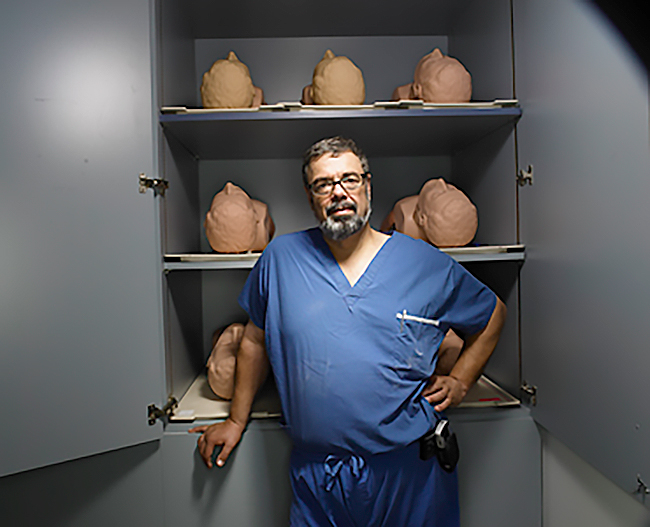 Professor Richard Reznick, wearing blue hospital scrubs, stands in front of an open cupboard containing sculptures of heads and torsos.