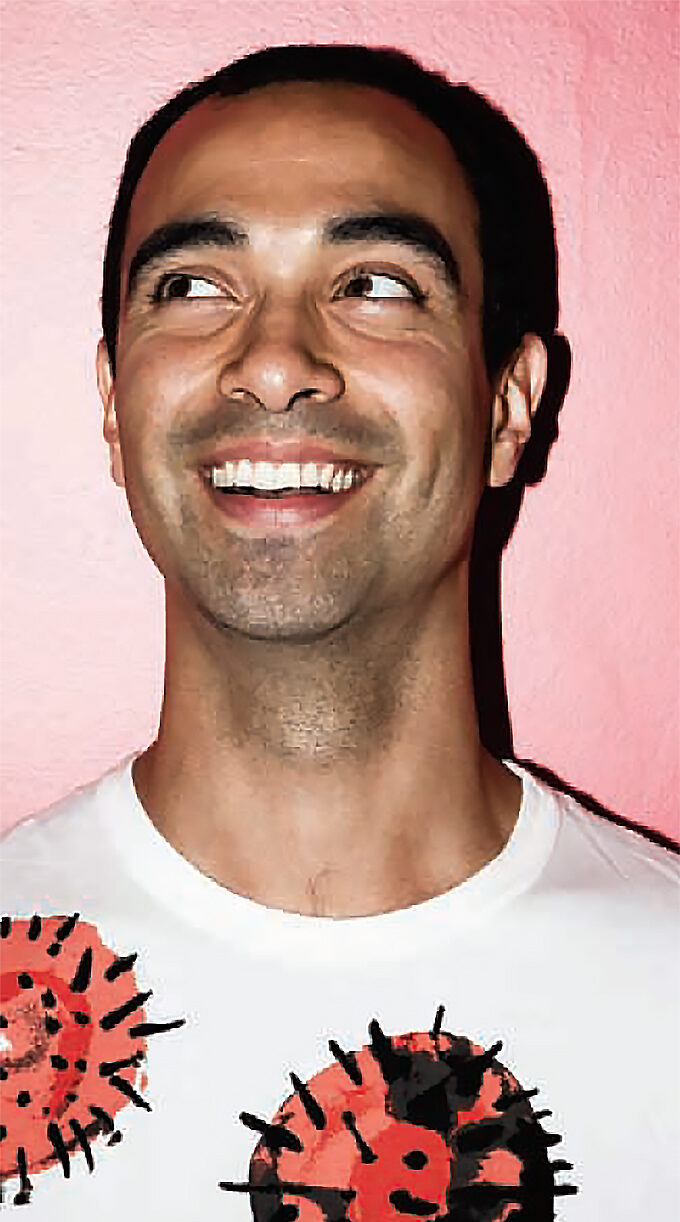 Luis Jacob in a white T-shirt with illustrations of orange and black circles with spikes, smiling and looking to his right