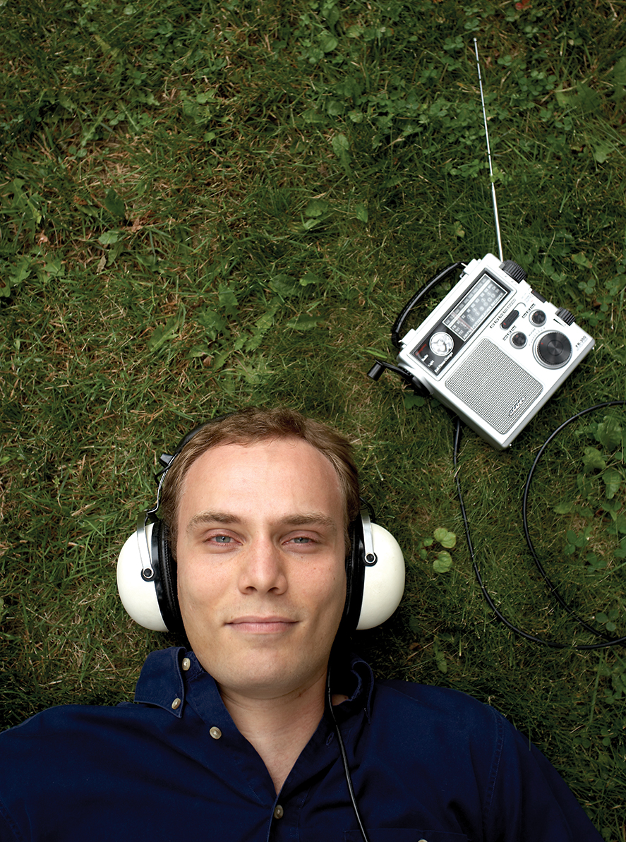 Bird's eye view of Jordan Poppenk, wearing white headphones, lying on a grassy field with a portable radio next to his head