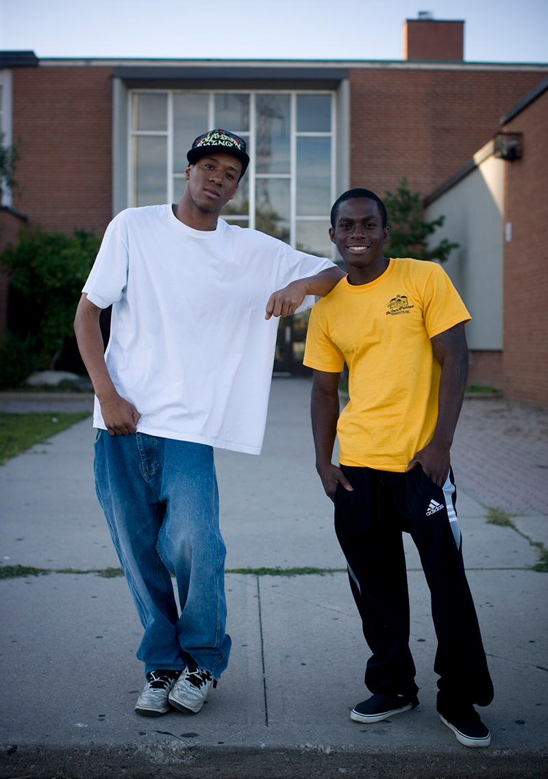 Omar Peters, in a white T-shirt, blue jeans, baseball cap and white sneakers, stands with an elbow resting on the shoulder of Prince Peprah, who is wearing a yellow T-shirt, black track pants and black sneakers. They are standing in front of a building with a panelled window and red-brown bricks.