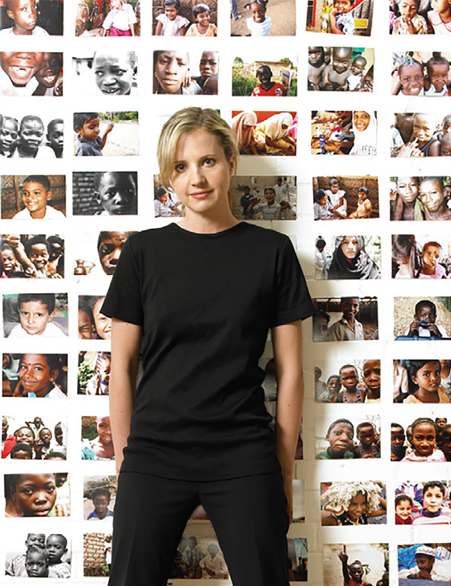 Samantha Nutt, wearing a black T-shirt and pants, stands in front of a wall filled with rows of photos of African children.