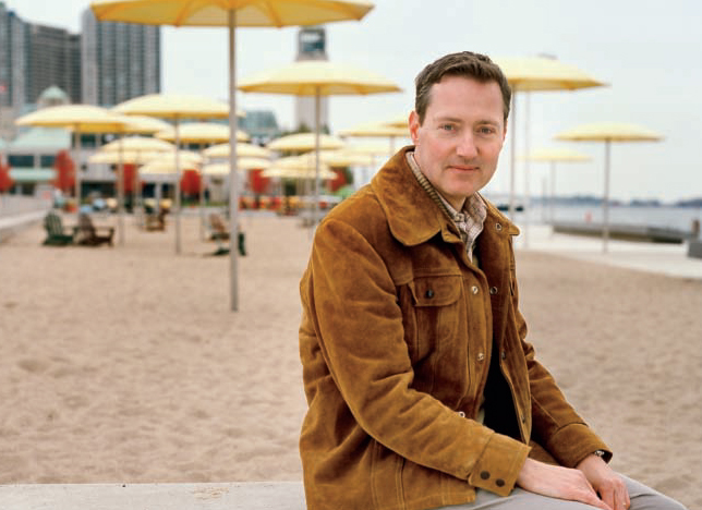 Mark Osbaldeston, sitting on a concrete surface, with yellow umbrellas on a sandy Toronto beach by the lakeshore in the background and condo buildings in the distance