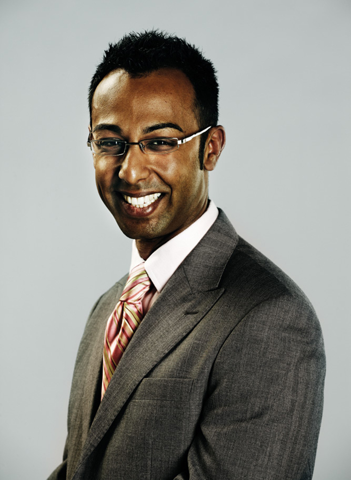 Headshot of Aly-Khan Rajani, smiling and wearing silver framed glasses, a red and olive green striped tie and a dark grey suit