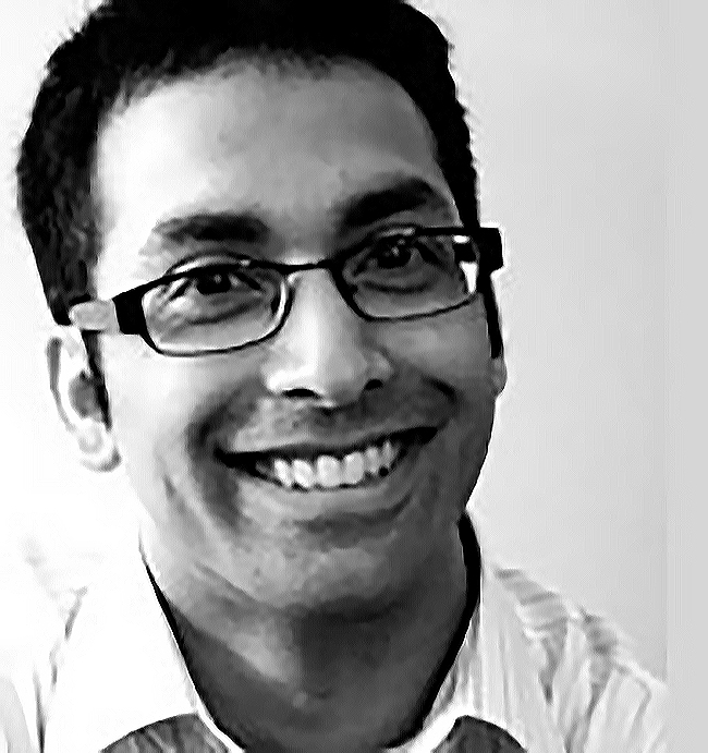 Black and white headshot of Andrew Pinto, wearing black framed glasses and a light-coloured, striped, collared shirt