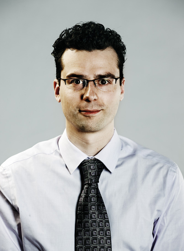 Headshot of Matthew Cimone, wearing black half-framed glasses, a light collared shirt and a black and grey patterned tie