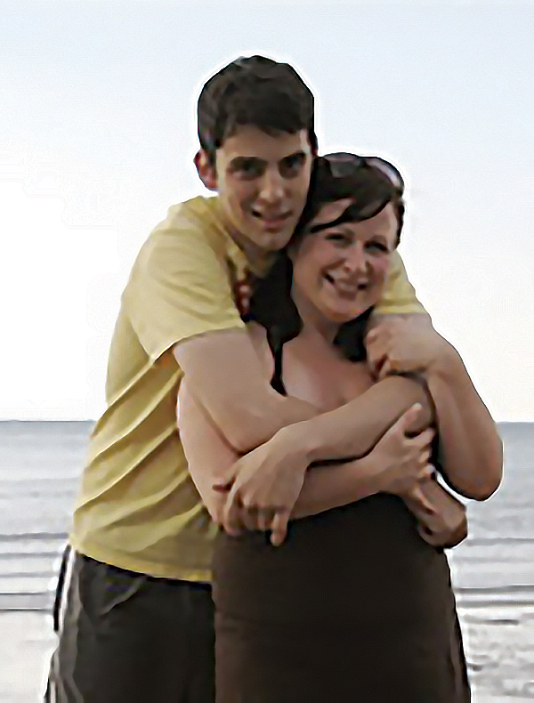 Brad Tapson, in a T-shirt and shorts, is standing behind Melanie Moore with his arms around her on a beach