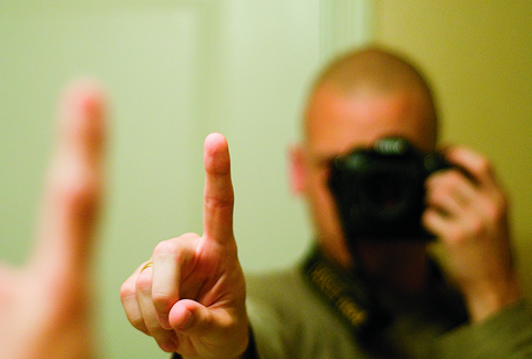 Photo of a man covering his face and holding up one finger into a mirror.