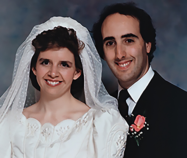 A studio headshot of Deborah Fletcher, wearing a white wedding gown and veil, and Wayne Levin in a black tie and jacket with a pink flower pinned to his lapel