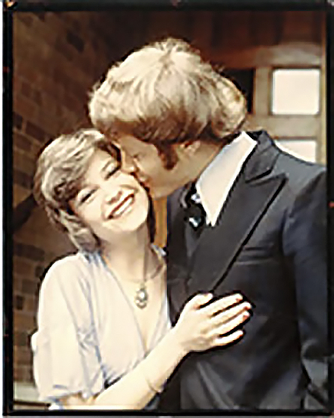 Outdoor shot of Monique in a light-coloured blouse or dress, smiling with one hand resting against Haijo, who is wearing a dark suit and tie and kissing her on the cheek