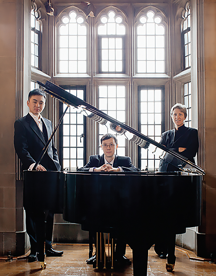 Kevin Lau sitting at a black grand piano in Hart House’s Music Room with Aaron Tsang and Prof. Gillian MacKay standing on either side.