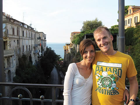 Trish De Luca and Ben Land are standing on an elevated bridge with metal railing. In the background are mid-rise buildings on either side surrounded by trees, a body of water in the far distance.