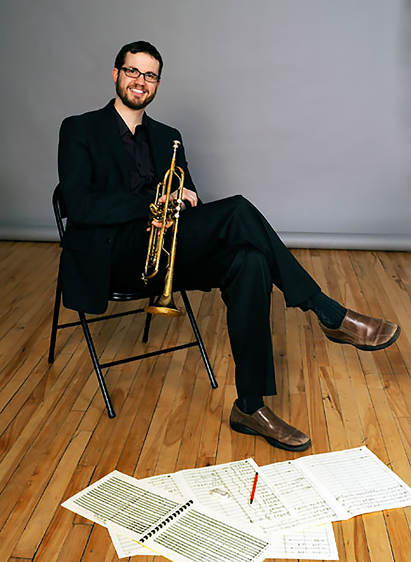Josh Grossman, dressed in a black suit and shirt and brown shoes, sits with his legs crossed in a metal chair, holding a trumpet. Sheets of music scores are scattered on the floor by his feet.