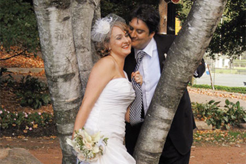Wedding photo of Tanya Koivusalo in a strapless wedding gown and white veil, holding a bouquet, and Adam Nayman in a suit, posing against the trunk of a tree, their cheeks nuzzling