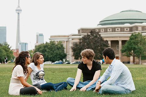 Four students sitting cross-legged on a grassy field at U of T's front campus, with Convocation Hall and CN Tower in the background
