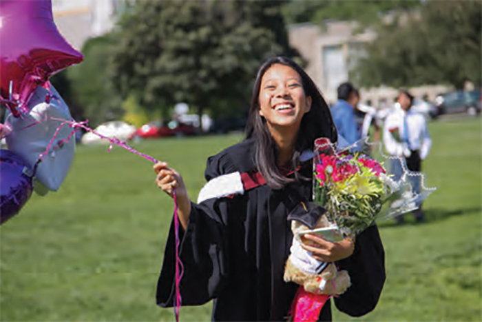 A student wearing convocation robes, standing on the field at King's College Circle, and holding balloons in one hand and a bouquet of flowers in the other