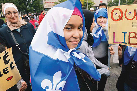 Photo of a woman wearing a hijab with a Quebecois flag draped over her.