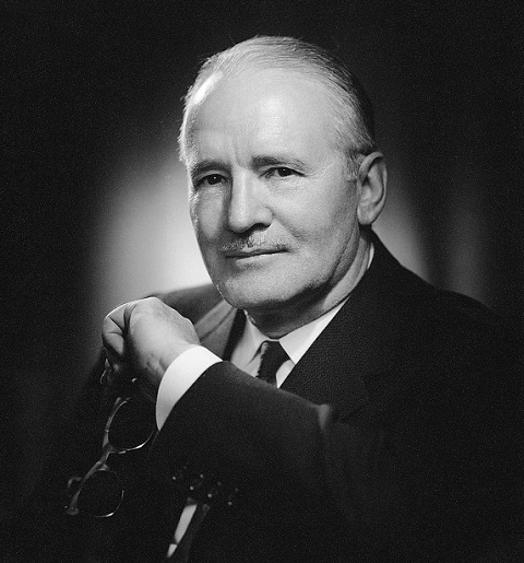 Black and white profile photo of Pay Bayly wearing a black suit and tie and holding his glasses