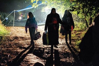 Photo of three refugees walking on a dirt path with their belongings at night