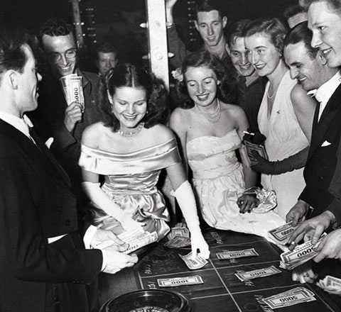Black and white photo of students in gowns and tuxedos placing bets around a roulette table