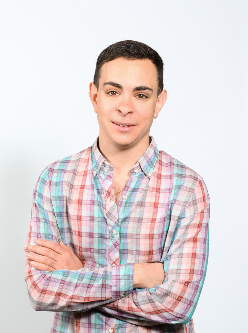 Studio shot of Ido Katri, wearing a light purple, cyan and red plaid, long-sleeved shirt, with arms crossed.