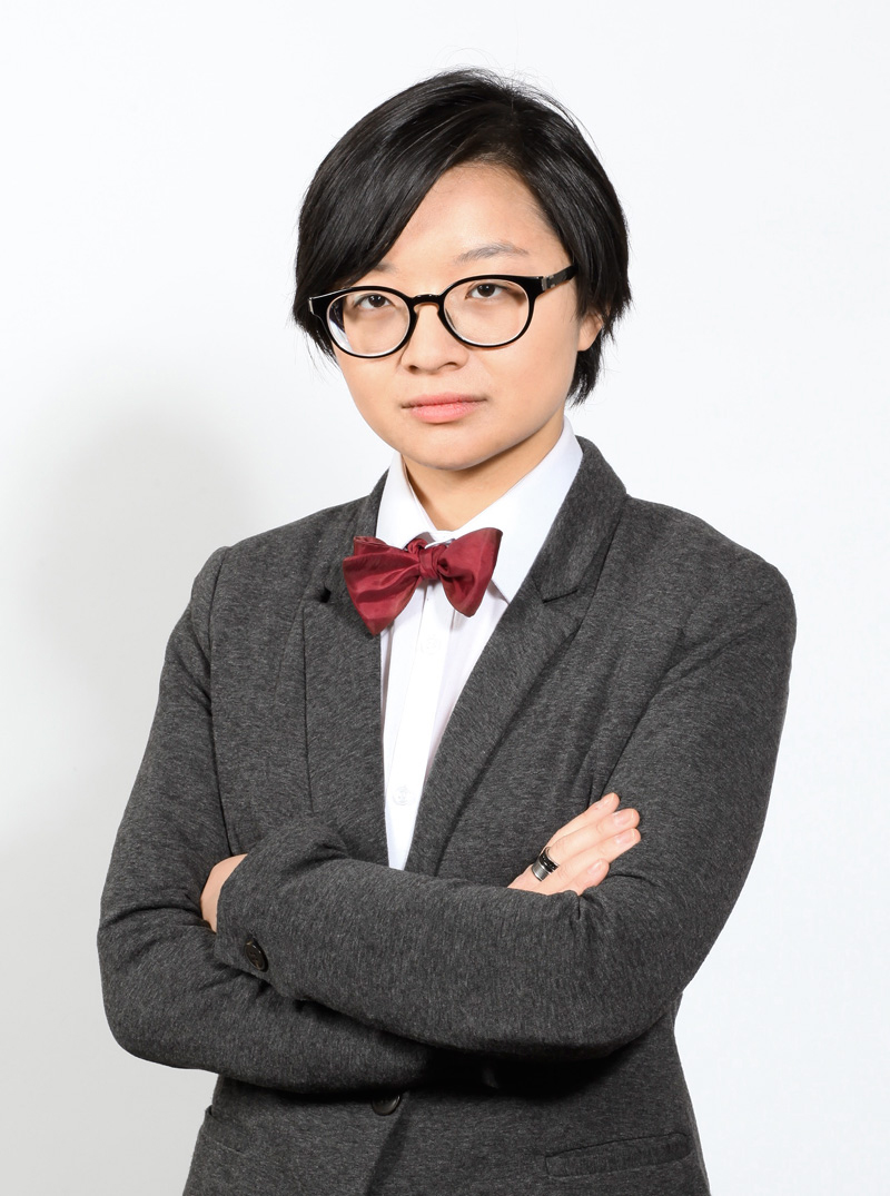 Studio shot of Iris Robin, wearing black-framed glasses, a white collared shirt under a dark grey wool blazer, and a red bow tie.