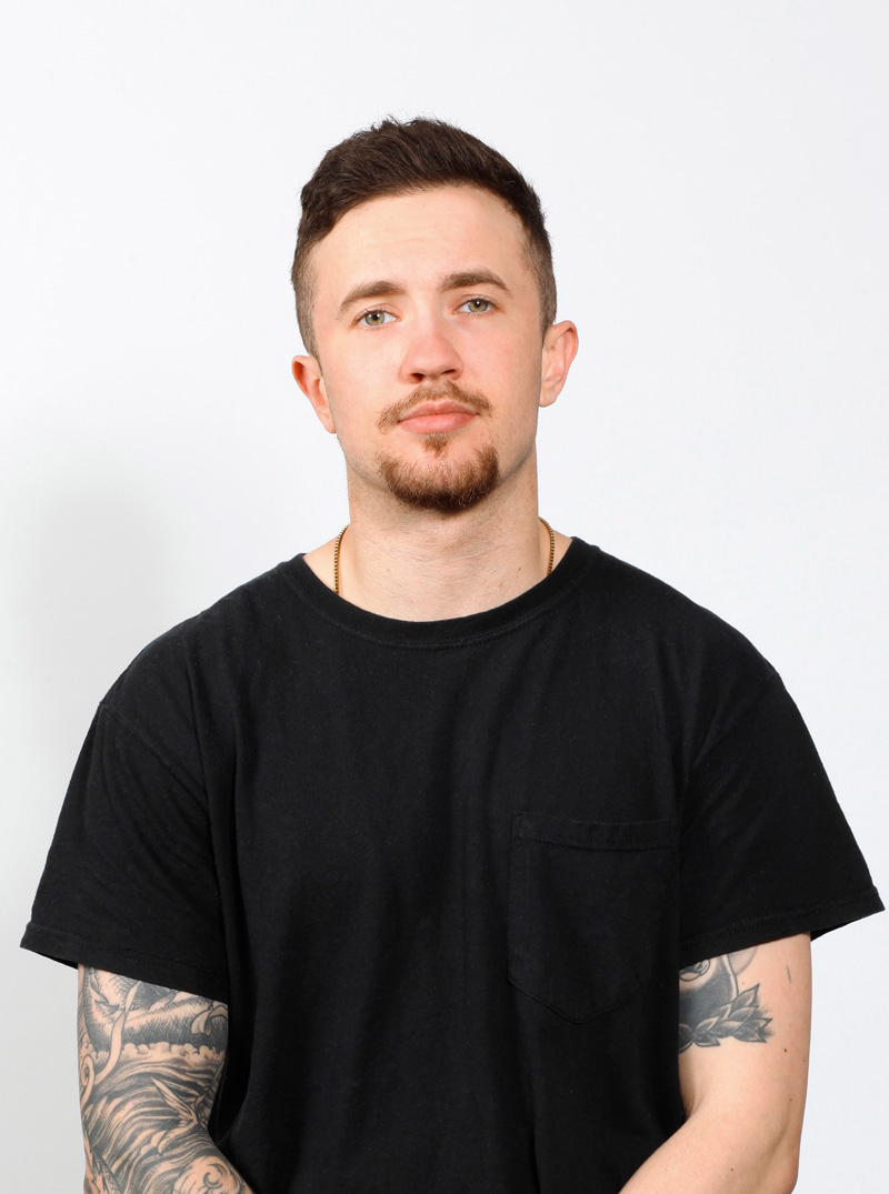 Studio shot of Seth Watts in a black T-shirt, with a goatee and tattoos on both arms.