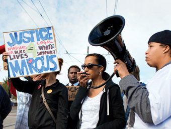 Photo of a group of protesters, one speaking through a loudspeaker and one holding a sign ("Justice 4 Marcus. Disregard for black lives = Legal white supremacy)