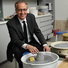 Kelvin Browne, wearing a dark grey suit and black tie, is seated in front of a potter wheel, one hand hovering over a grey lump of clay in the centre of the wheel