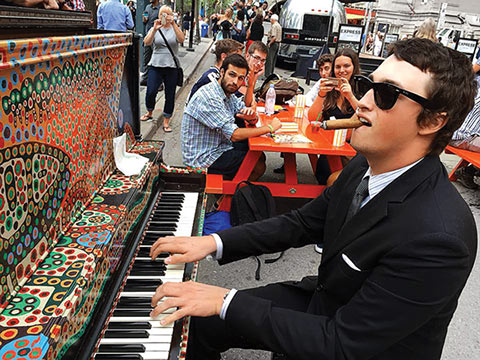Photo of Sebastian Brown with sunglasses and a cigar in mouth, playing piano on the street, and onlookers at a picnic bench