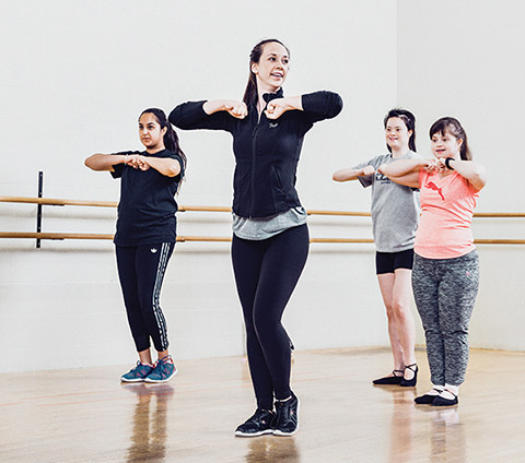 Photo of Neha Singh and Jade Ryan teaching a dance move to two young dancers.