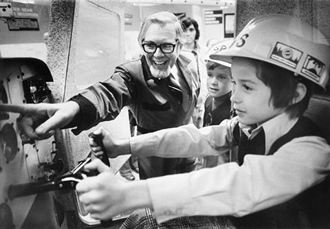 Bernard Etkin pointing at a feature, speaking to two boys wearing hard hats, one of whom is steering the simulator.