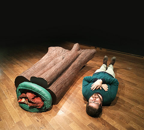 Photo of an art installation of a sleeping bag inside a log on the left and a man lying with eyes closed on the right.