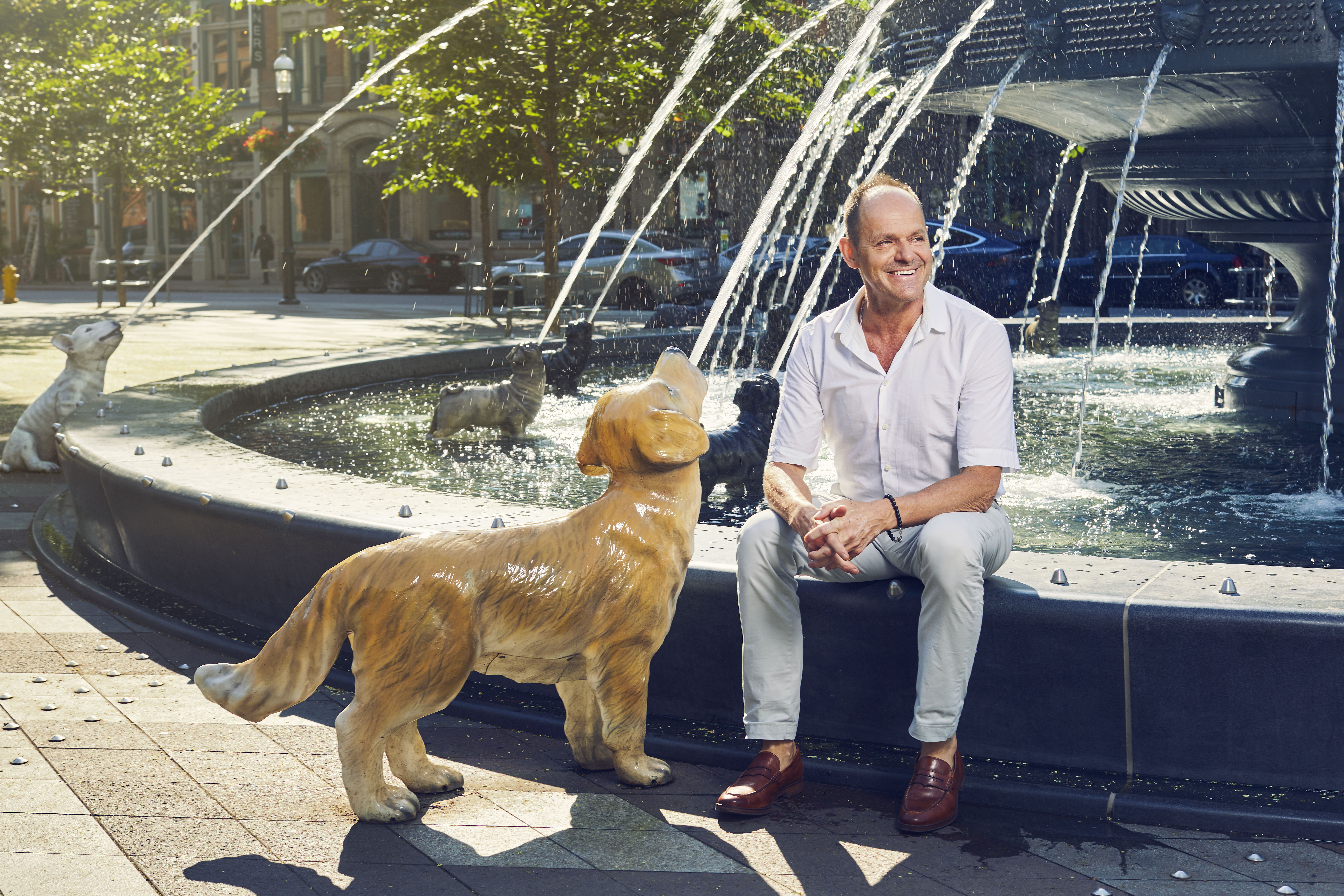 Claude Cormier sitting on edge of dog fountain he designed. He is beside a cast-iron golden retriever