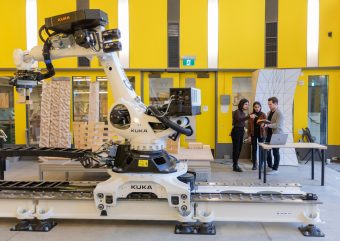 White robotic arm in the foreground against a yellow wall. Three students engaged in conversation in the background