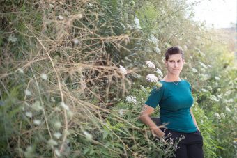 Prof. Jessica Green in a turquoise top standing in a clearing next to a forest of bushes, branches and white flowers