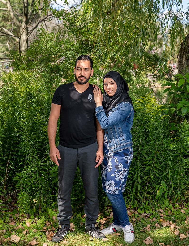 Alaa Al Saleh and Hanaa Al Bitar are standing in front of green bushes under leaves from a tree, looking at the camera. Bitar is turned toward Saleh with her hands on his left shoulder.