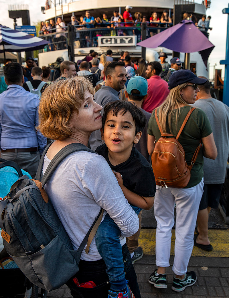 Jennifer Nagel is wearing a backpack and carrying Mohamad Al Saleh, who is smiling at the camera, while standing in line for the ferry. In the background, a ferry with passengers onboard is in dock.