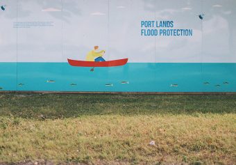 Photo of a drawing of a person rowing a boat next to the words "Port Lands Flood Protection" on a wall erected on grass