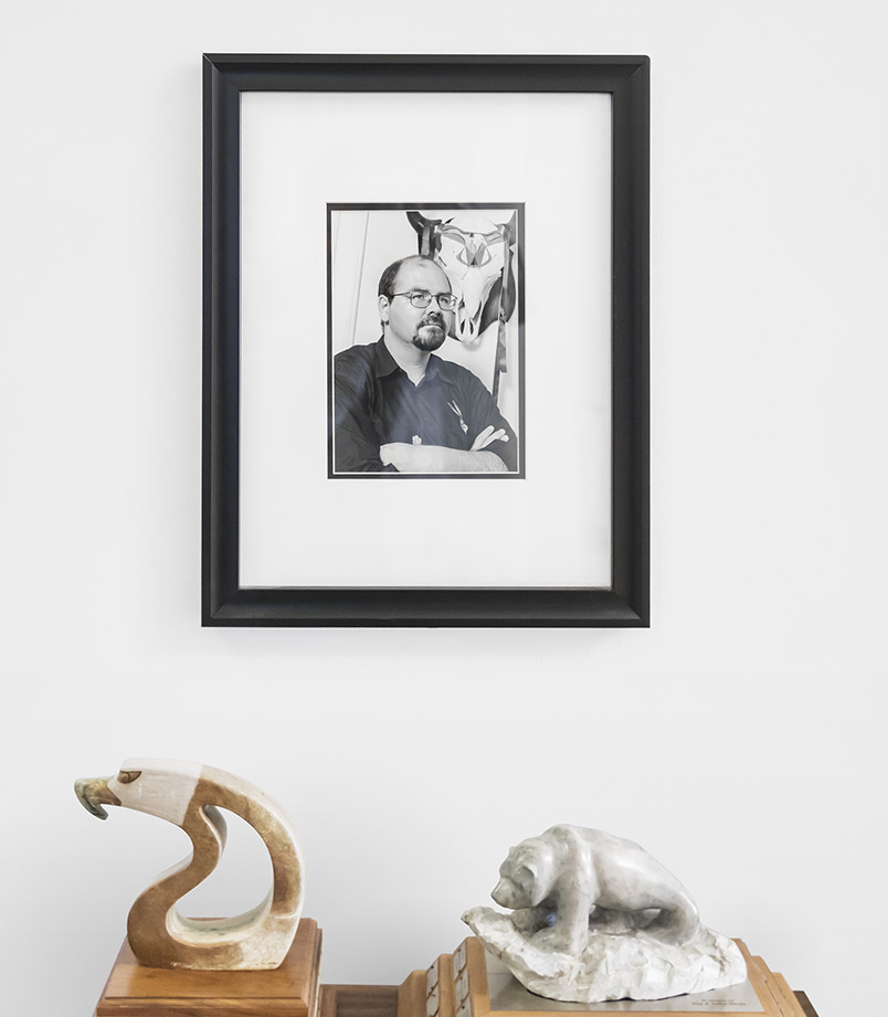 A framed black and white photo of Rodney Bobiwash hanging on a white wall above two awards, one a stone carving of an eagle and the other a stone carving of a grizzly bear on a rock, both on wooden plaques