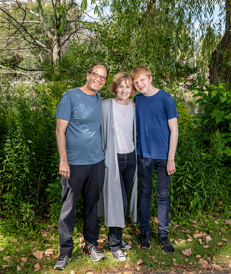 Sergio Tenenbaum, Jennifer Nagel and Leo Tenenbaum are smiling at the camera and standing in front of green bushes. In the background are overhanging leaves from a tree.