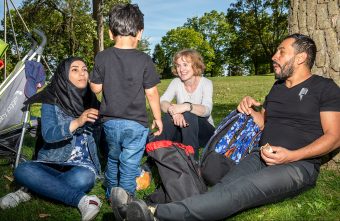 Hanaa Al Bitar, Jennifer Nagel and Alaa Al Saleh are sitting in a grassy field on Centre Island, looking at Mohamad Al Saleh, who is standing. Nagel is smiling and Saleh is leaning against a tree.