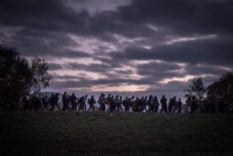 Migrants walking in line in the distance across a grassy field in the early morning or evening