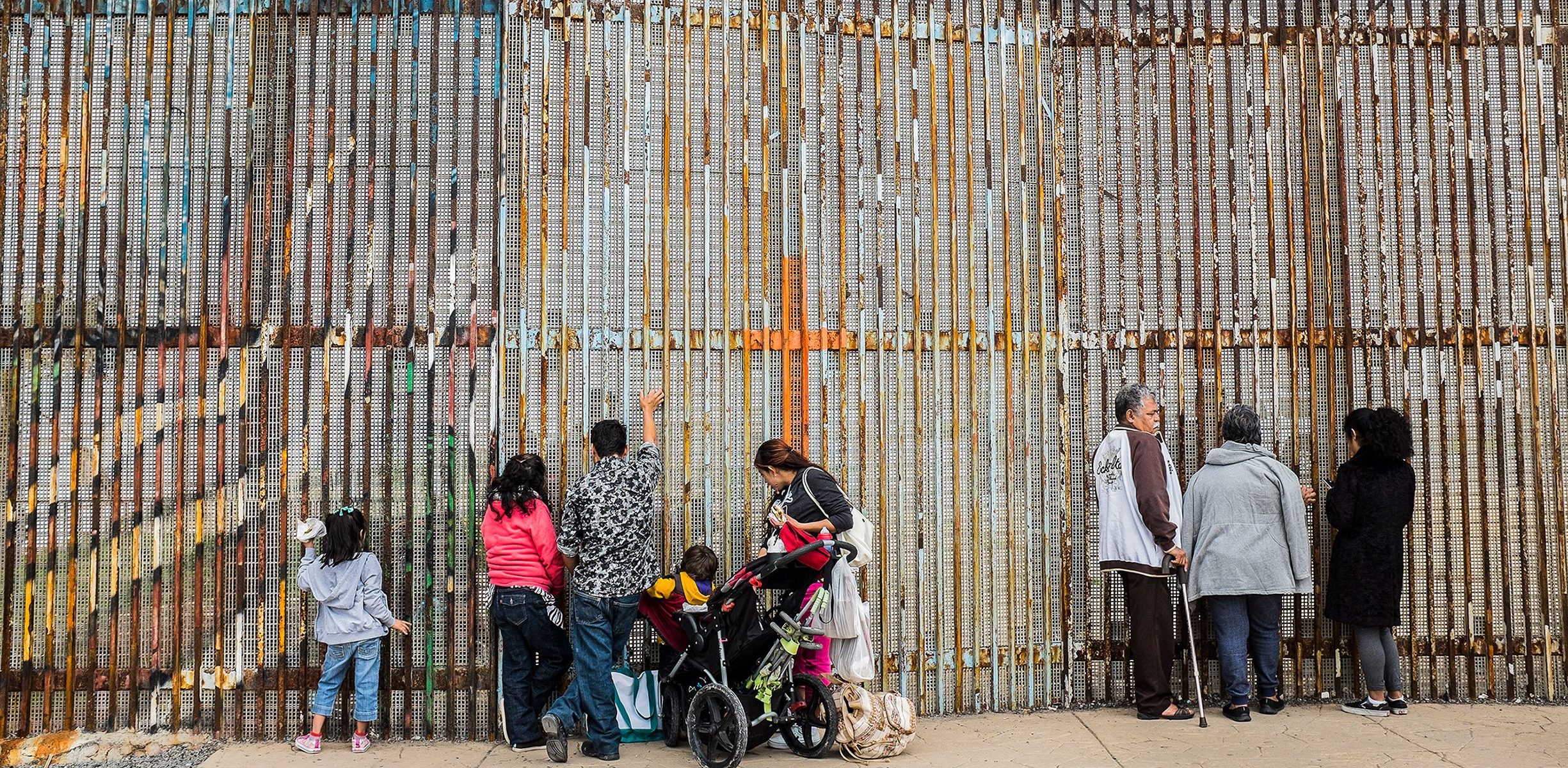 In the foreground, a family of five, with two young girls and a toddler in a stroller, stand facing the border gates in the background. An elderly couple and another adult stand a few feet away also facing the gates.