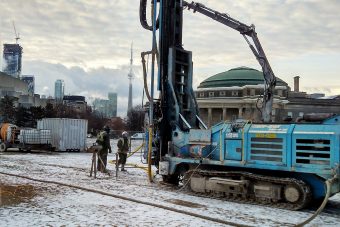 Two workers standing in front of a tank-like machine drilling a hole into the ground of the front campus field