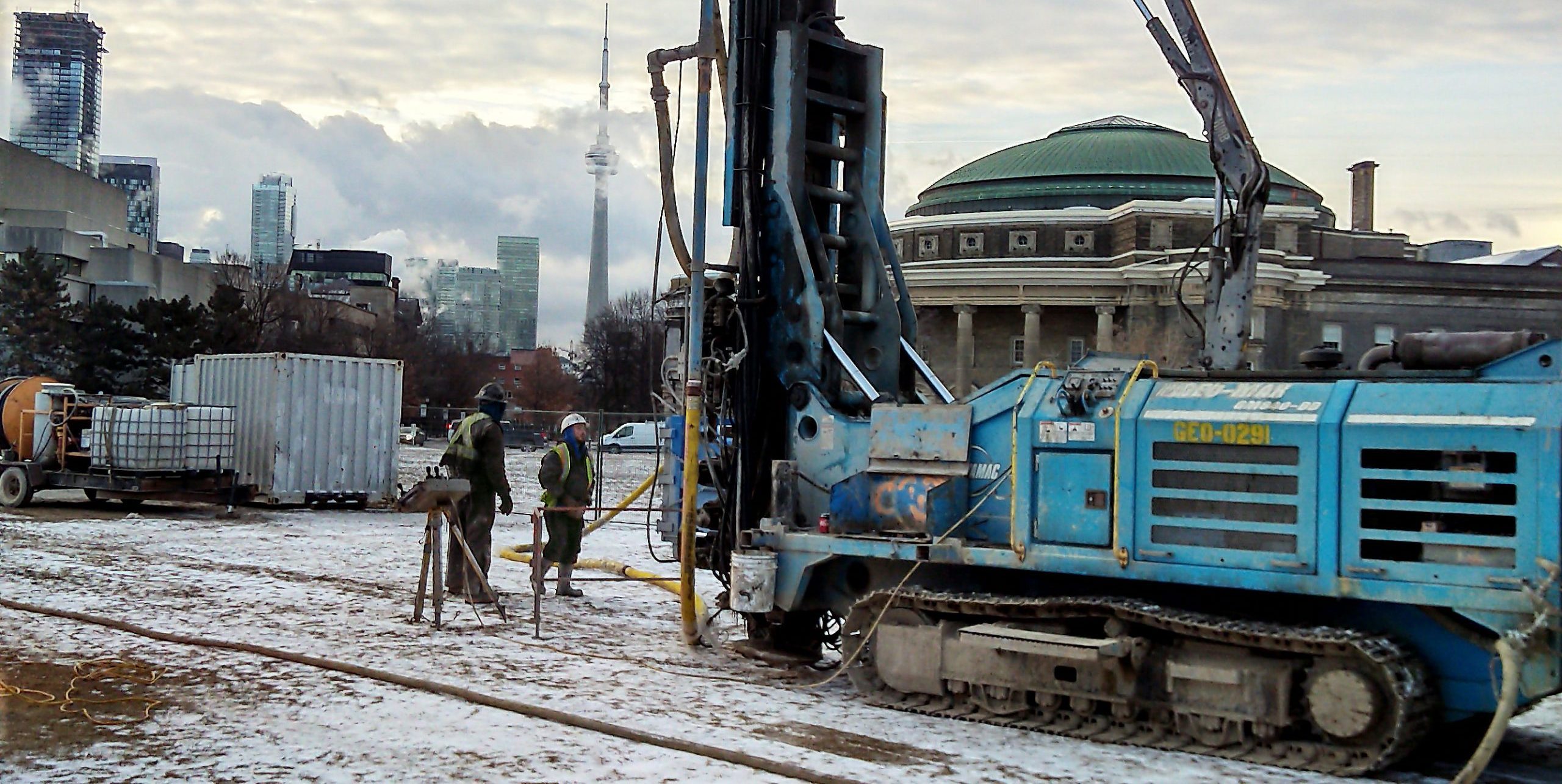 Two workers standing in front of a tank-like machine drilling a hole into the ground of the front campus field